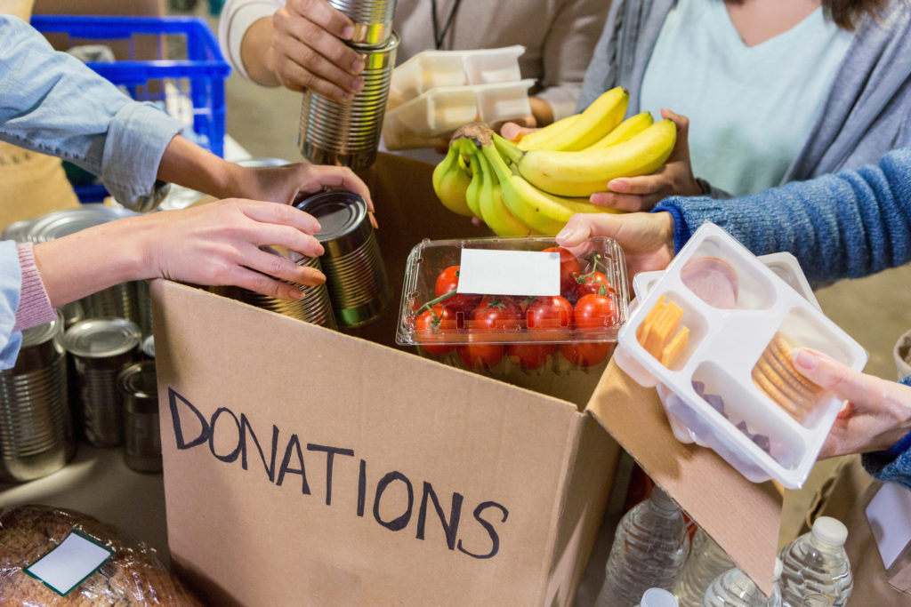 A group of unrecognizable food bank volunteers stand around a table and unpack food items from a cardboard box labeled, "Donations".
